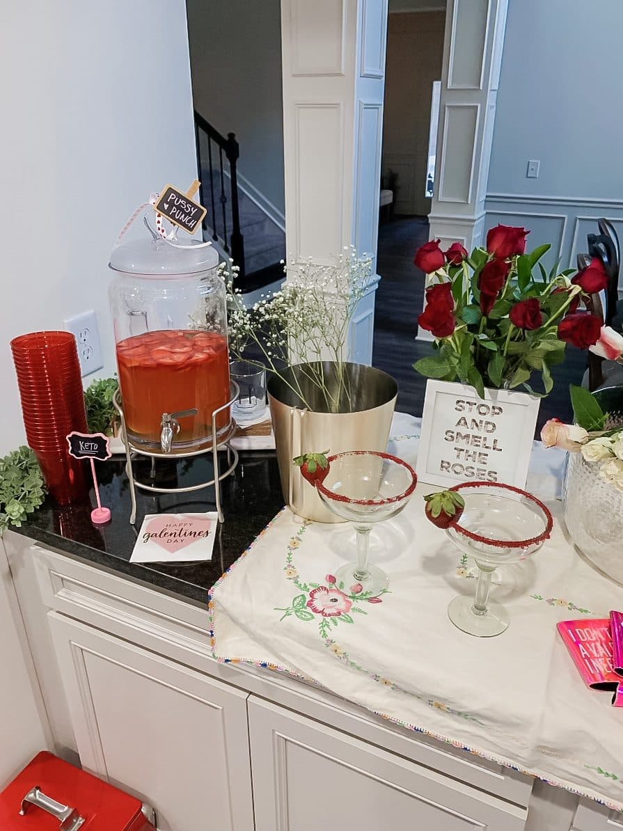 A drinks bar with a punch dispenser, strawberry-rimmed margarita glasses, red roses, and a 'Stop and Smell the Roses' sign.