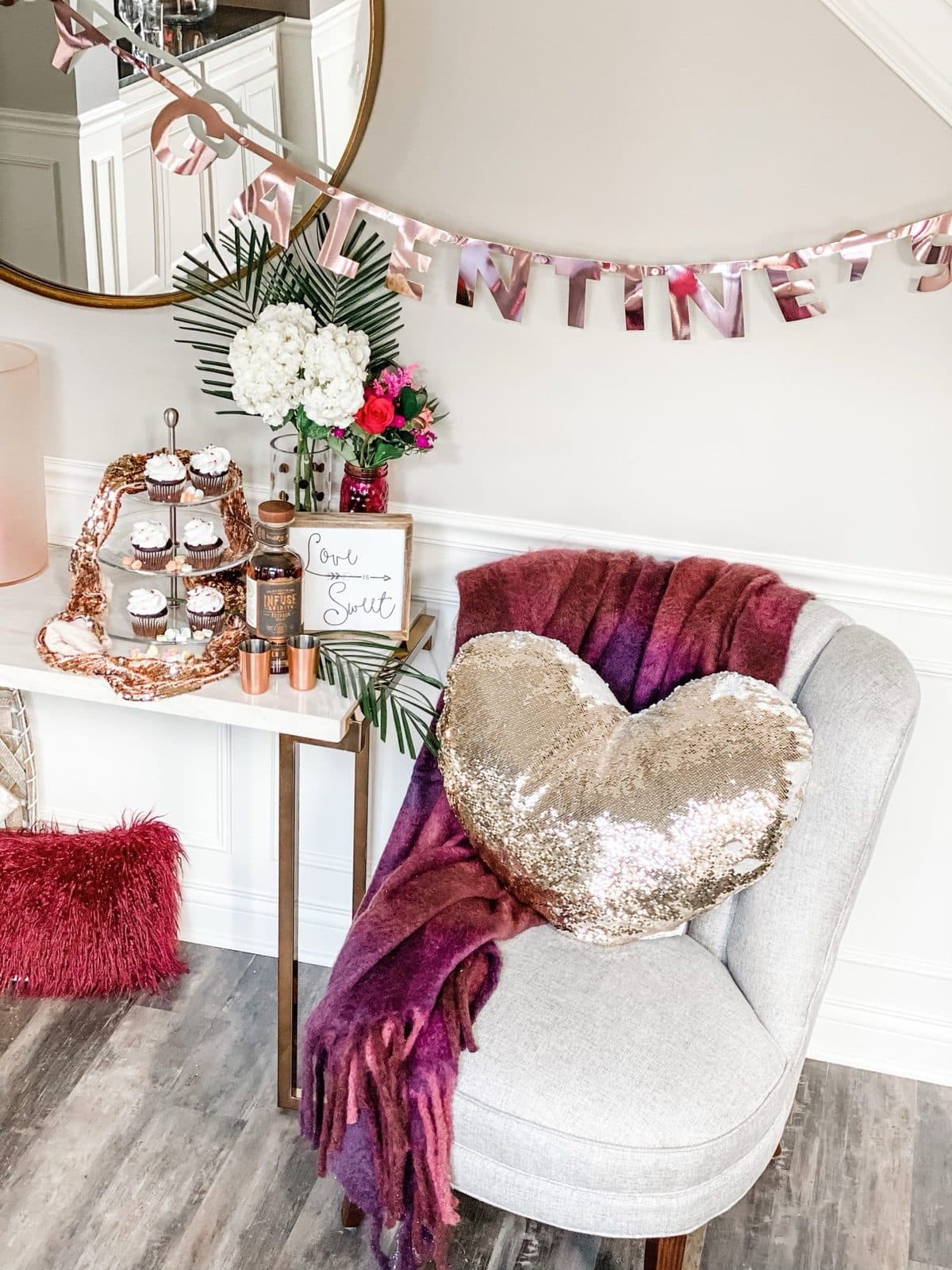A lounge vignette, a gold sequin heart pillow and plum throw on a grey chair beside a marble console with a cupcake tier and a 'Love Spout' sign under a rose-gold 'Galentine's' banner.