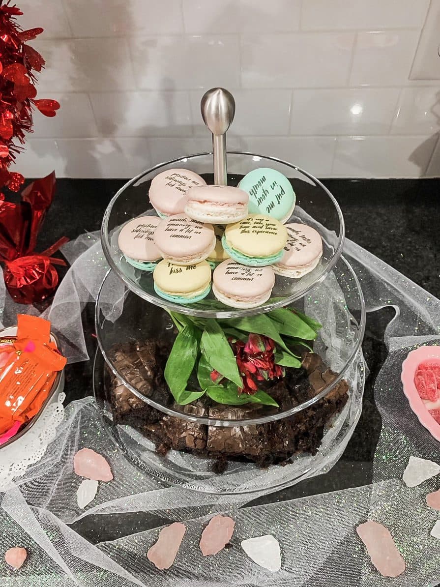 A tiered stand of pastel macarons printed with messages, over a brownie base on a glittered cloth scattered with rose quartz.