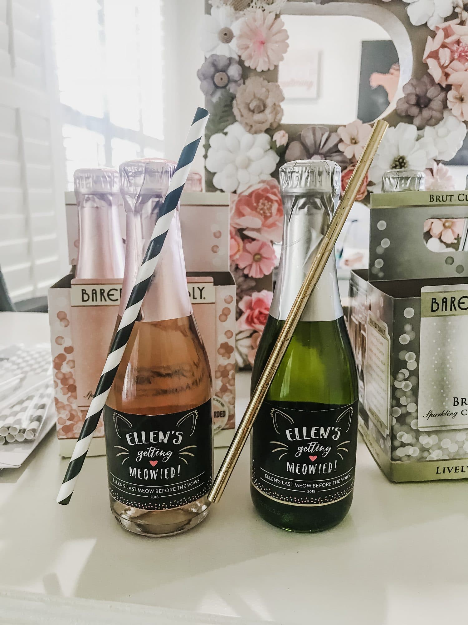 Mini champagne bottles labeled 'Ellen's getting Meowied!' with striped straws, in front of a floral paper backdrop.