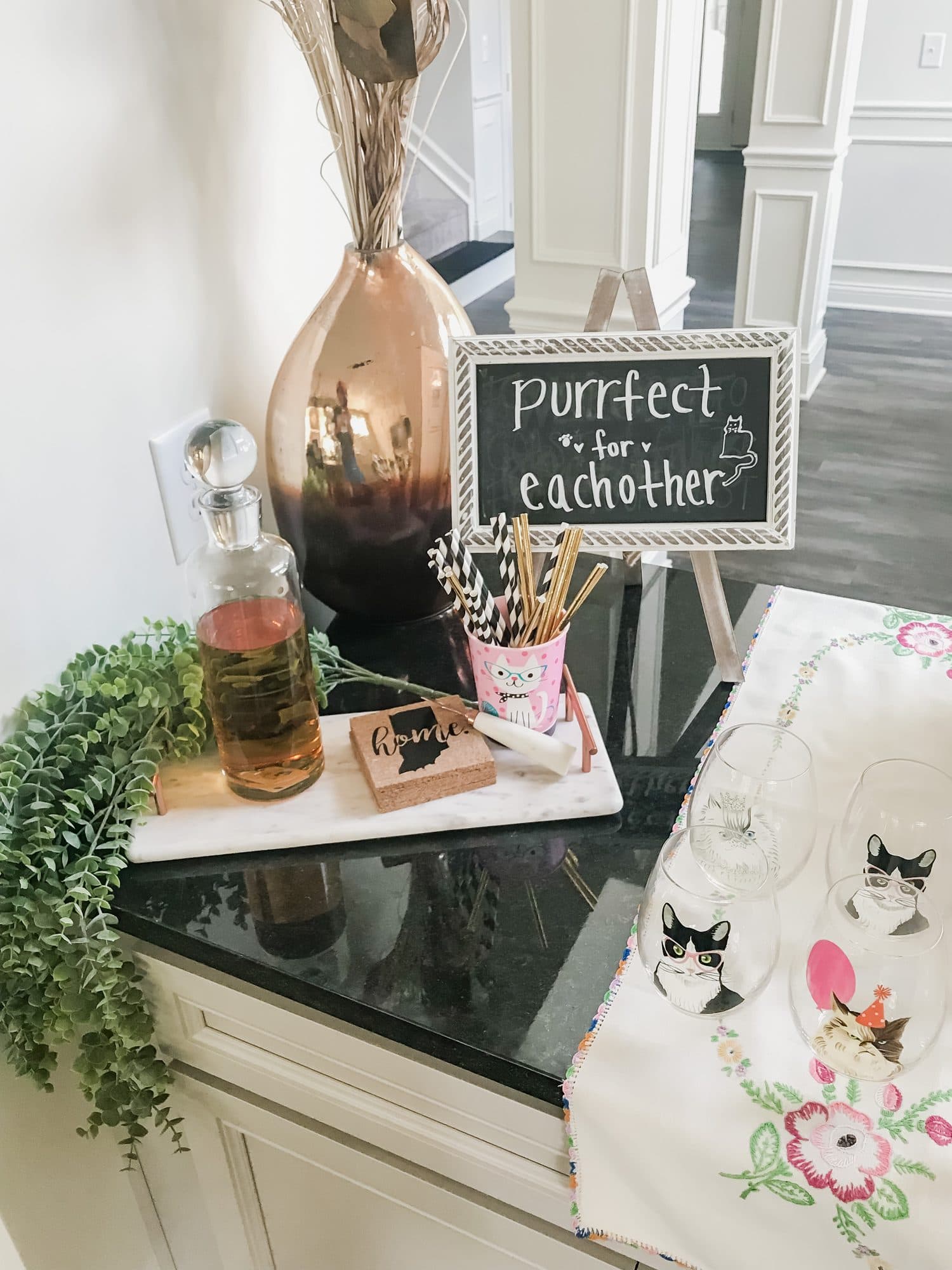 A styled bar vignette, a copper vase and whiskey decanter on a marble tray with a 'purrfect for each other' chalk sign and cat-face glasses.