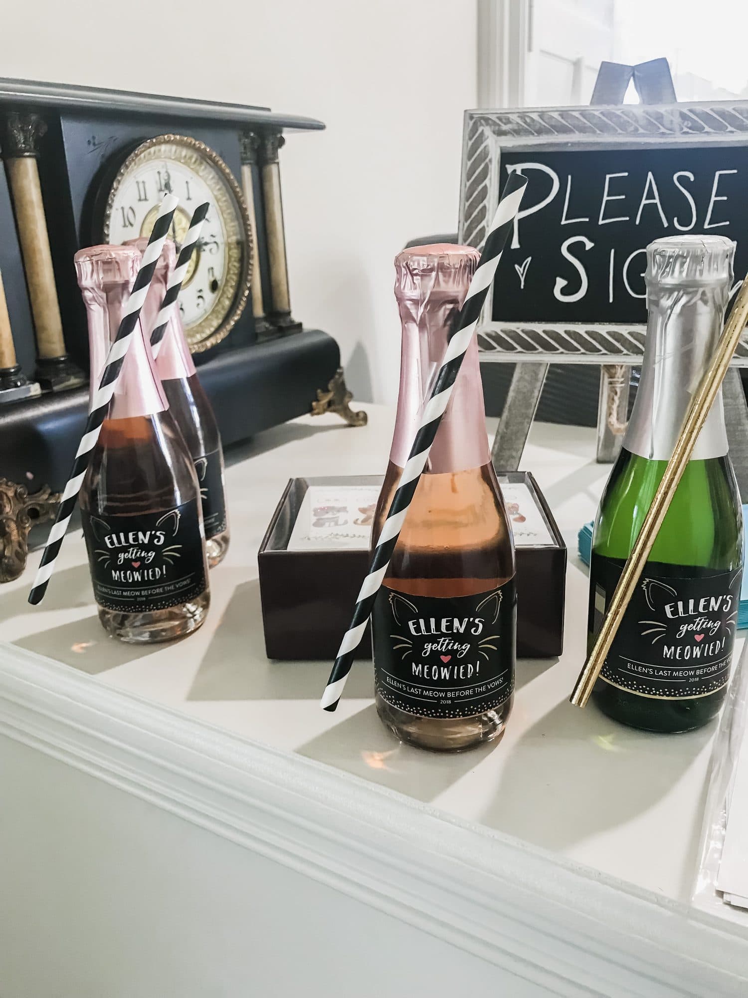A favor-and-guestbook station, rosé mini-bottles with gold straws lined up by a 'Please Sign' easel and an antique mantel clock.