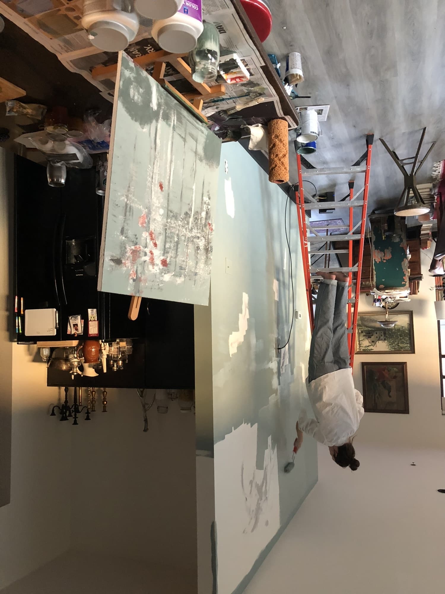 Haley on a ladder hand-painting a soft blue sky-and-cloud mural across a large interior wall, paint cans and rollers staged below.