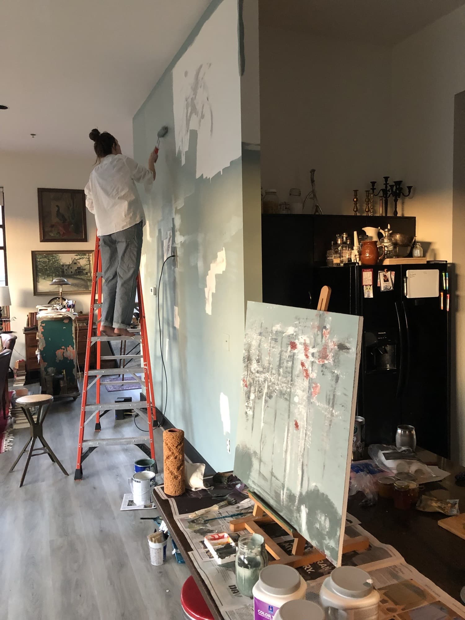 Haley on a ladder hand-painting a soft blue sky-and-cloud mural across a large interior wall, paint cans and rollers staged below.