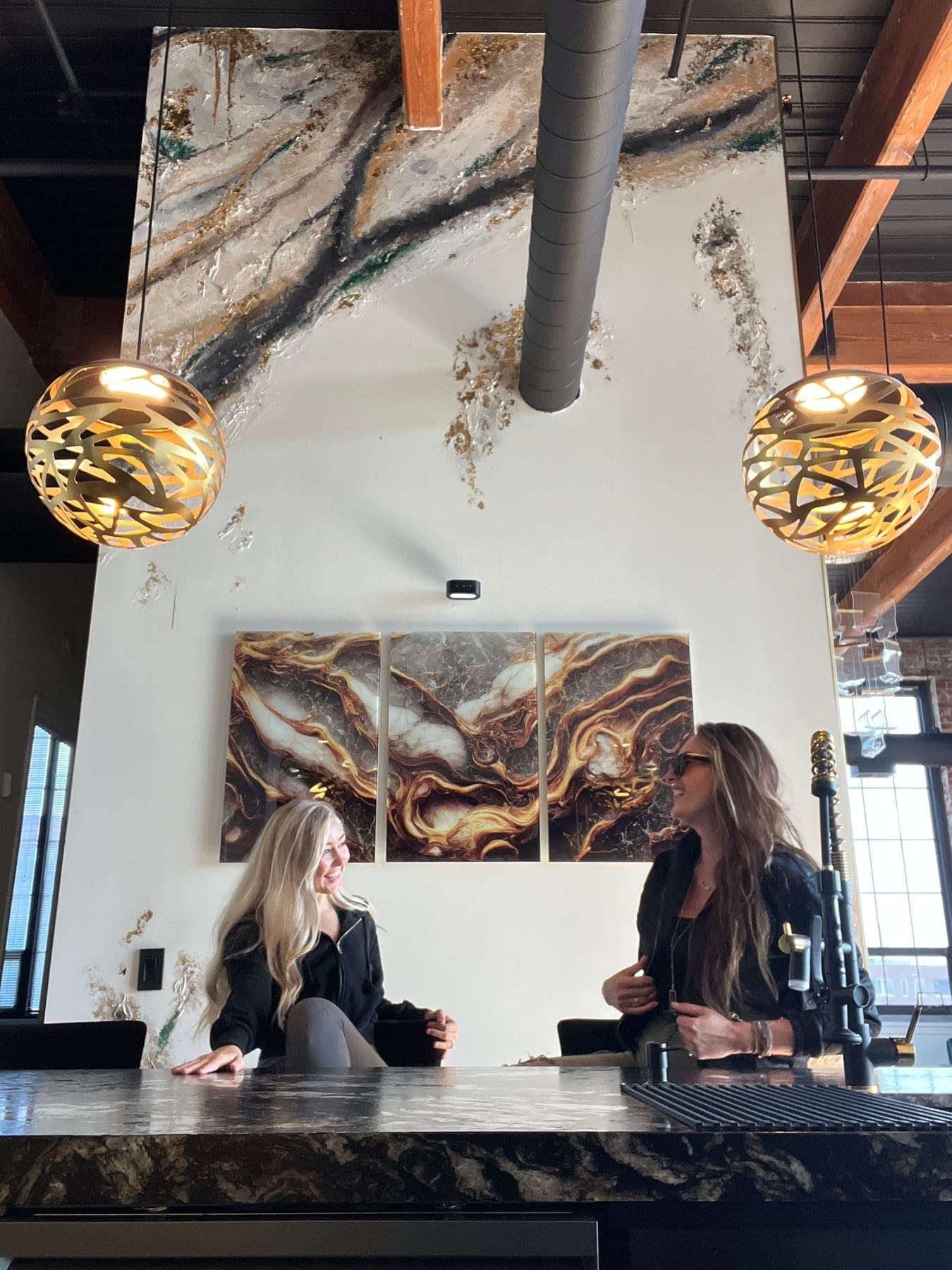 Two women talking across the black-marble bar under gold pendant lights, the poured-gold triptych and the feature wall behind them.