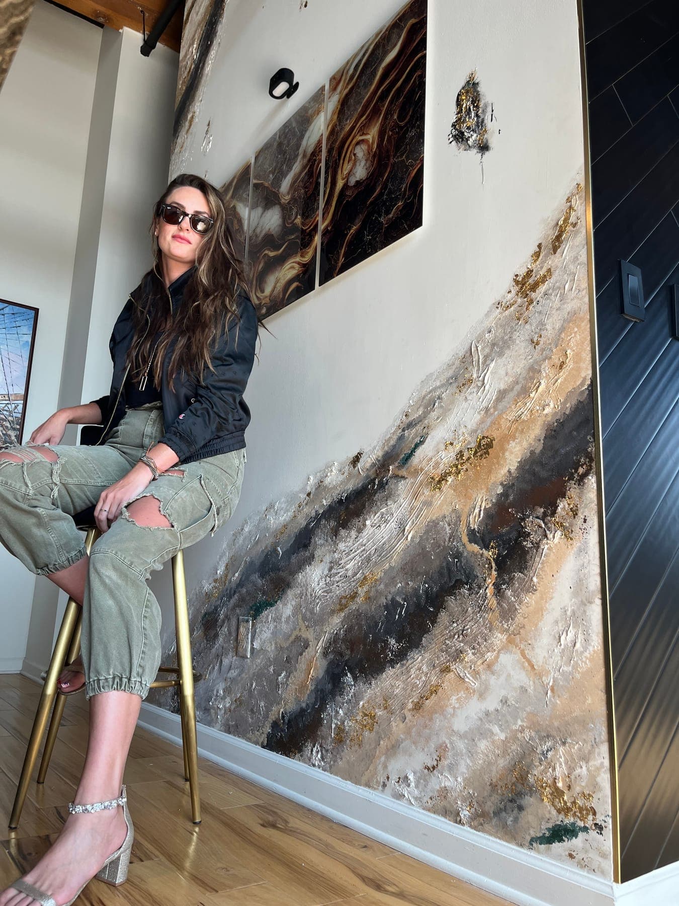 A woman in sunglasses on a brass stool beside the feature wall, the gold-veined slab and inset art rising behind her.