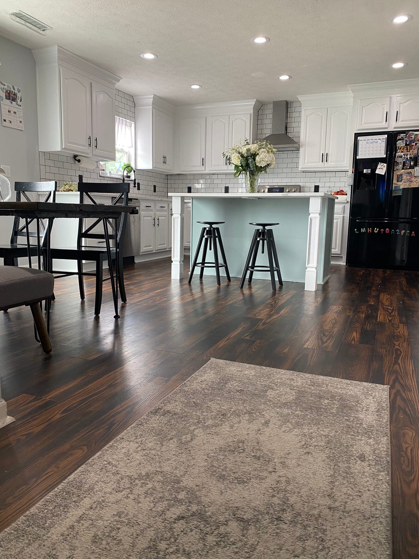 The whole kitchen from the dining side: white cabinets and subway tile, a sage-green island with black metal stools and white hydrangeas, dark wood floors under a faded gray rug.