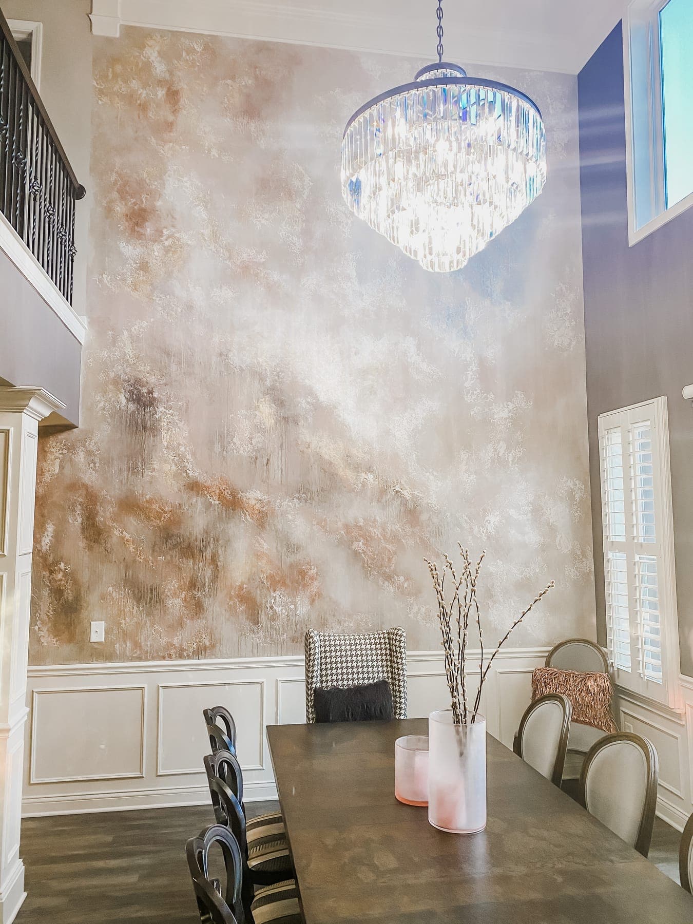 The full two-story feature wall, hand-finished copper and pewter plaster rising past the balcony rail to a crystal drum chandelier, pussy willows on the table below.