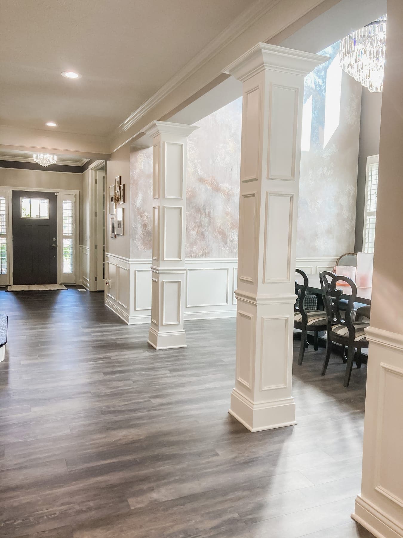 The dining room from the entry, white paneled columns and wainscoting framing the plaster wall, the front door and gallery frames down the hall.