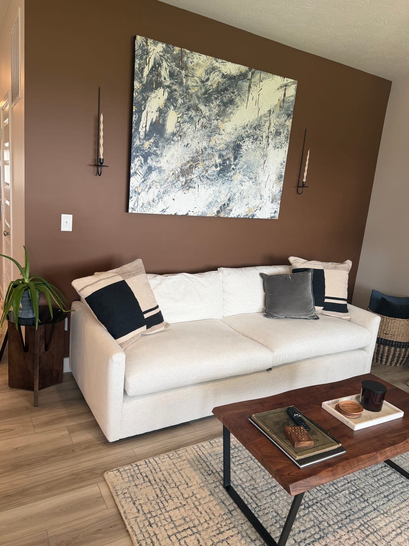 The living room's rust accent wall with a large abstract canvas between two black sconces, a white sofa with black and gray pillows, and a walnut coffee table.