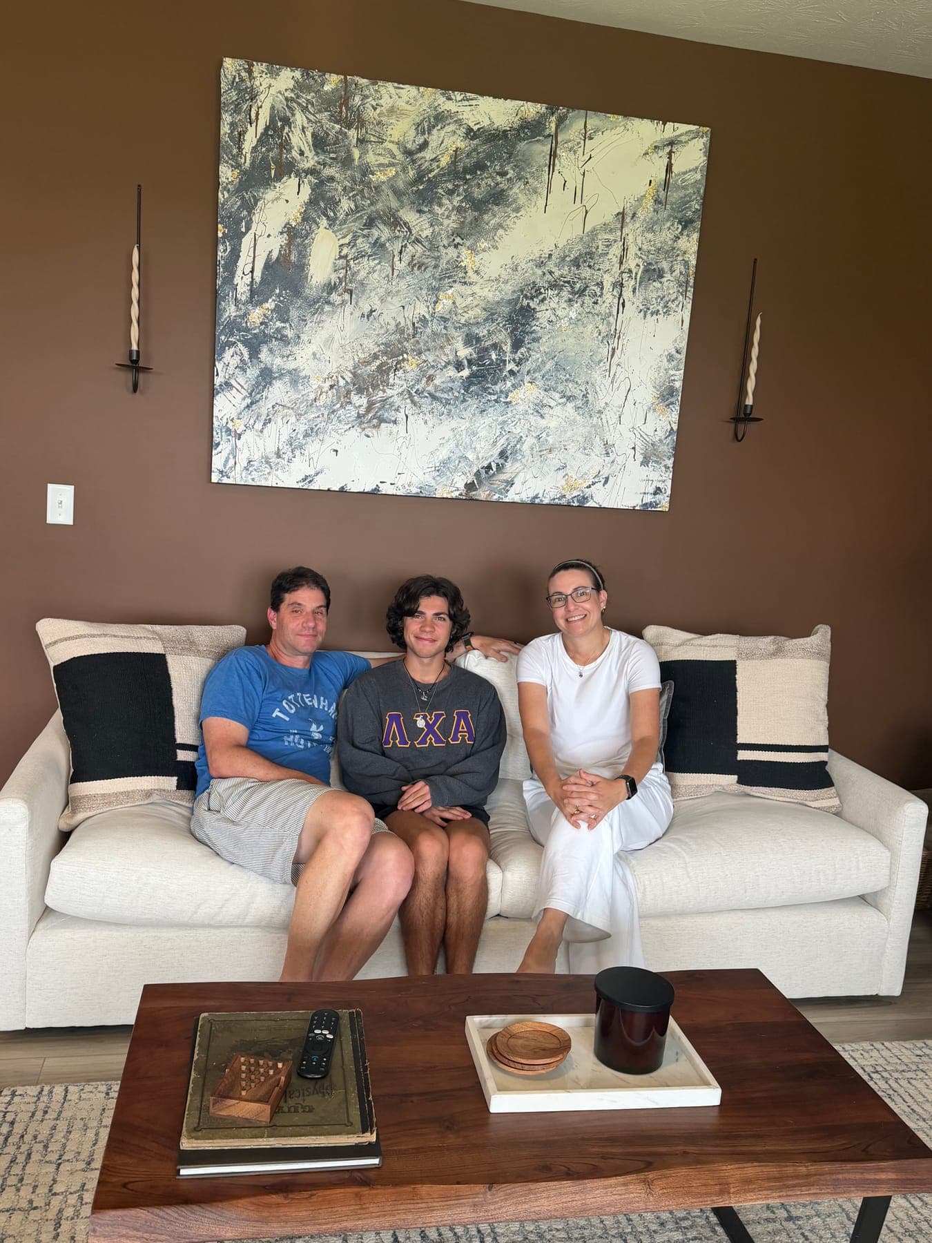 The family of three on the white sofa under the abstract canvas, styled books and a wood tray on the coffee table in front of them.