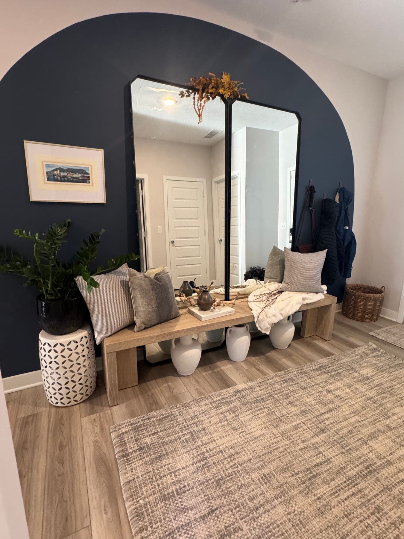 The entry, a navy painted arch framing a tall black-framed mirror, a pale wood bench with layered pillows and a throw, white vases and a woven basket below.