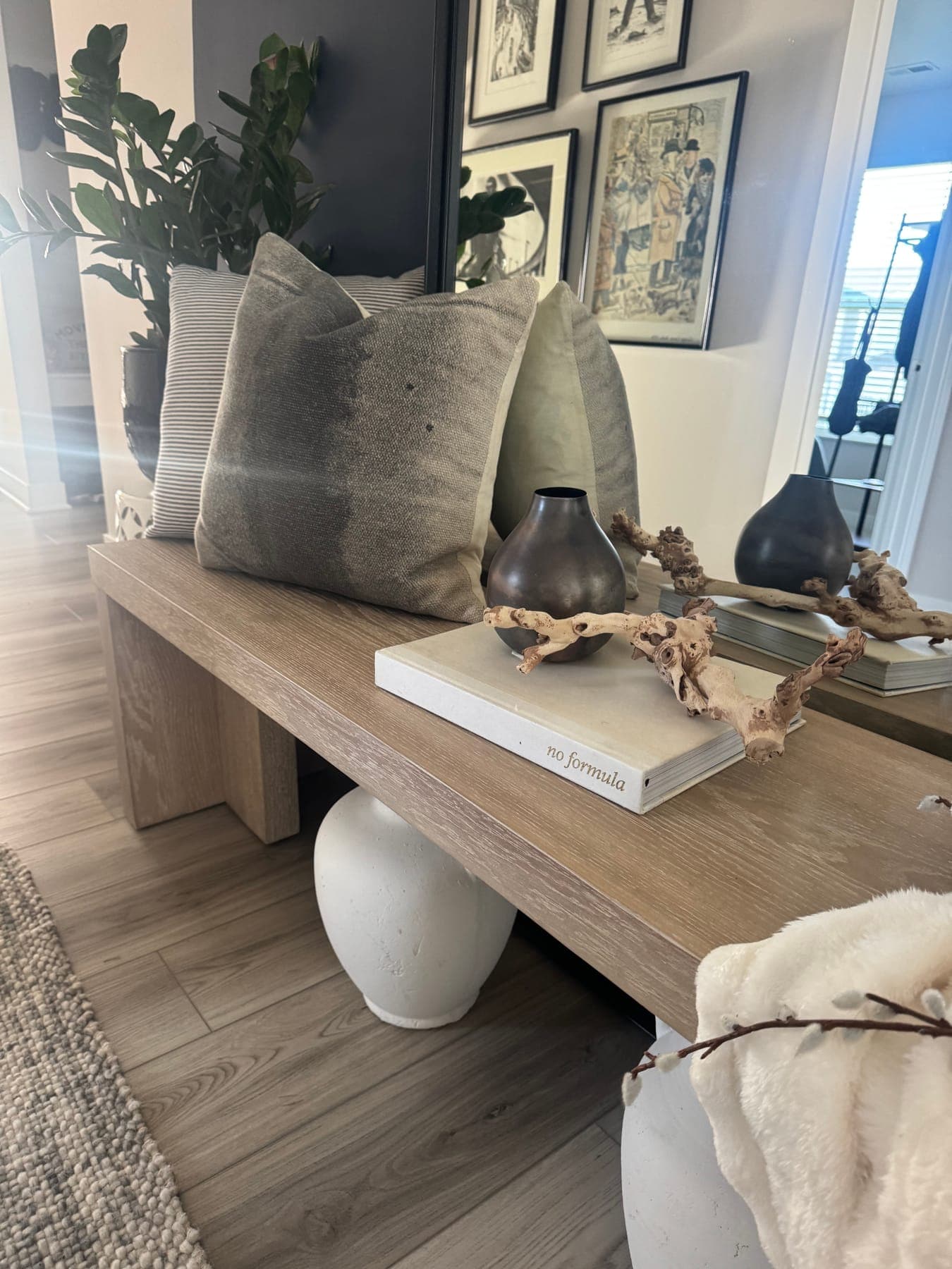 Detail of the entry bench: gray and cream pillows, a bronze vase, a book, and dried stems on the pale wood seat.