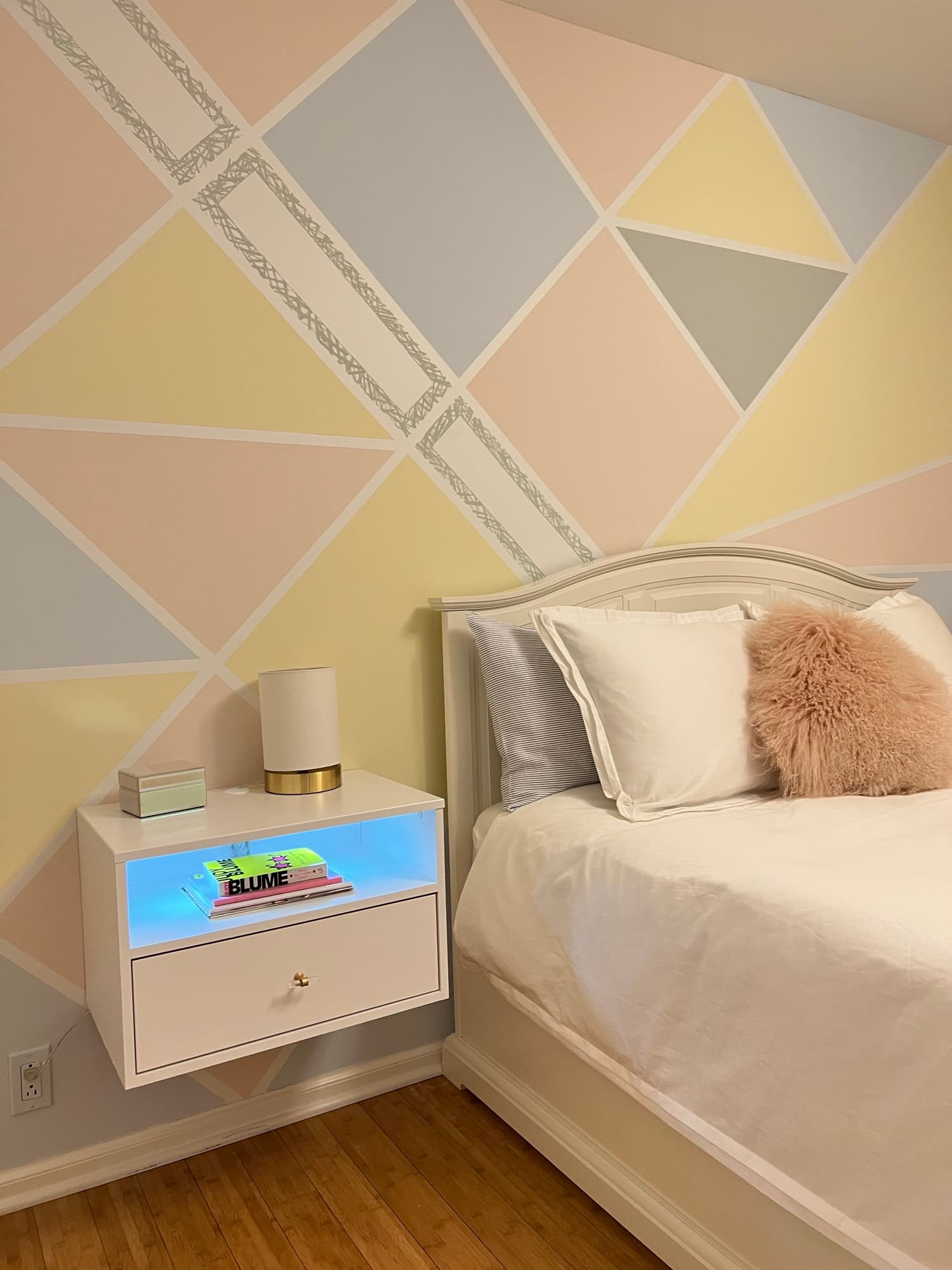 Close on the bed, blush faux-fur and striped pillows against the diamond wall, a floating nightstand glowing blue beneath a brass lamp.
