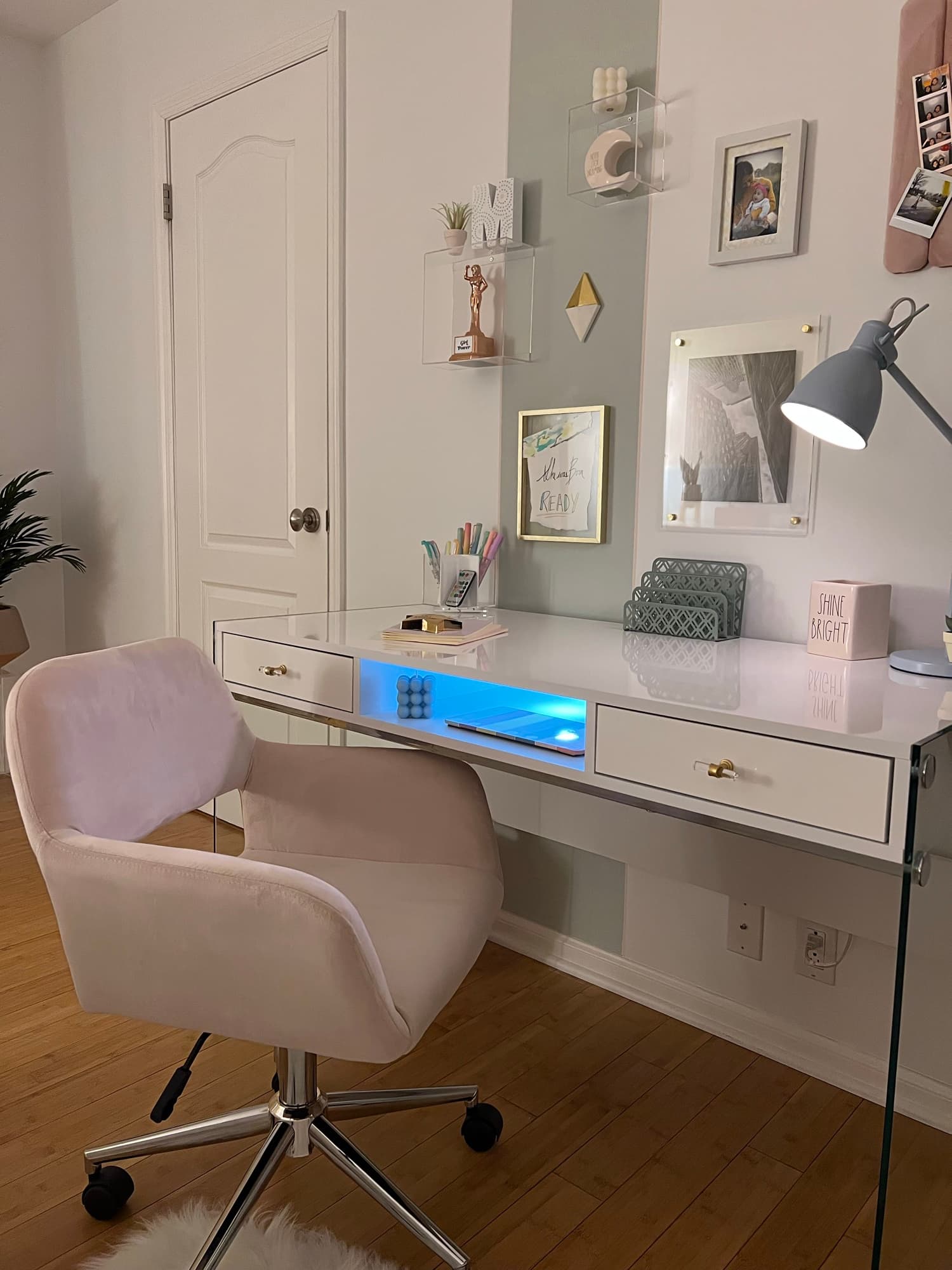 A glossy white vanity desk with a mirrored back panel, gold accents, a blush velvet chair, and 'Shine Bright' styling boxes.