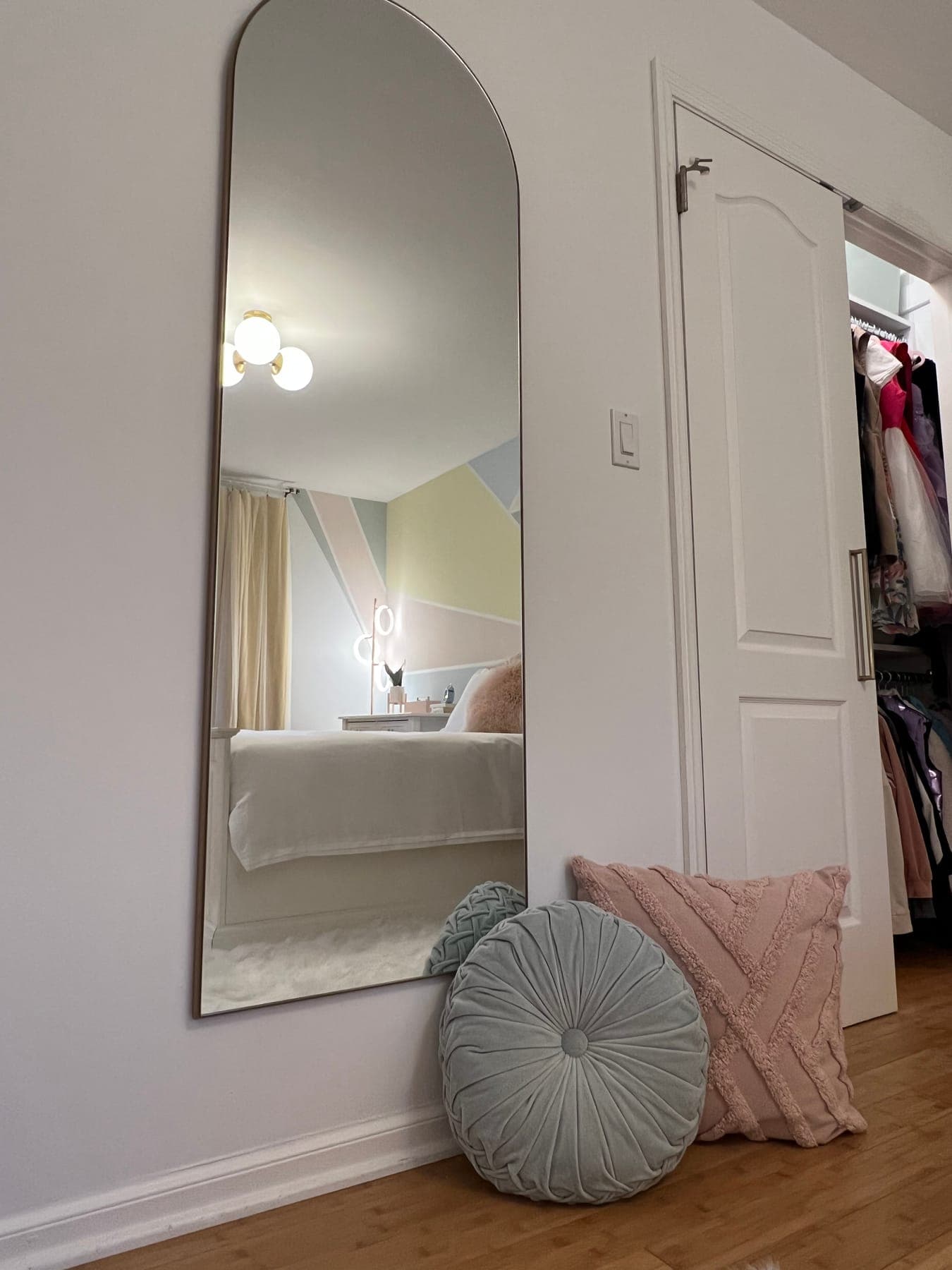 A full-length arched mirror by the closet door reflecting the diamond wall and bed, a sage pleated pouf and blush pillow on the floor beneath it.