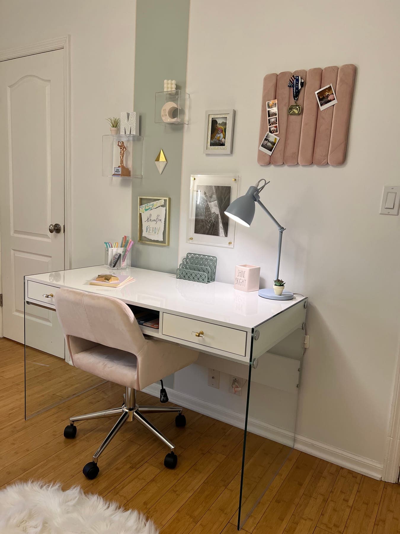 A white desk on clear acrylic legs with a blush velvet chair, a sage lamp, a photo ledge, and a pink pegboard on the wall.