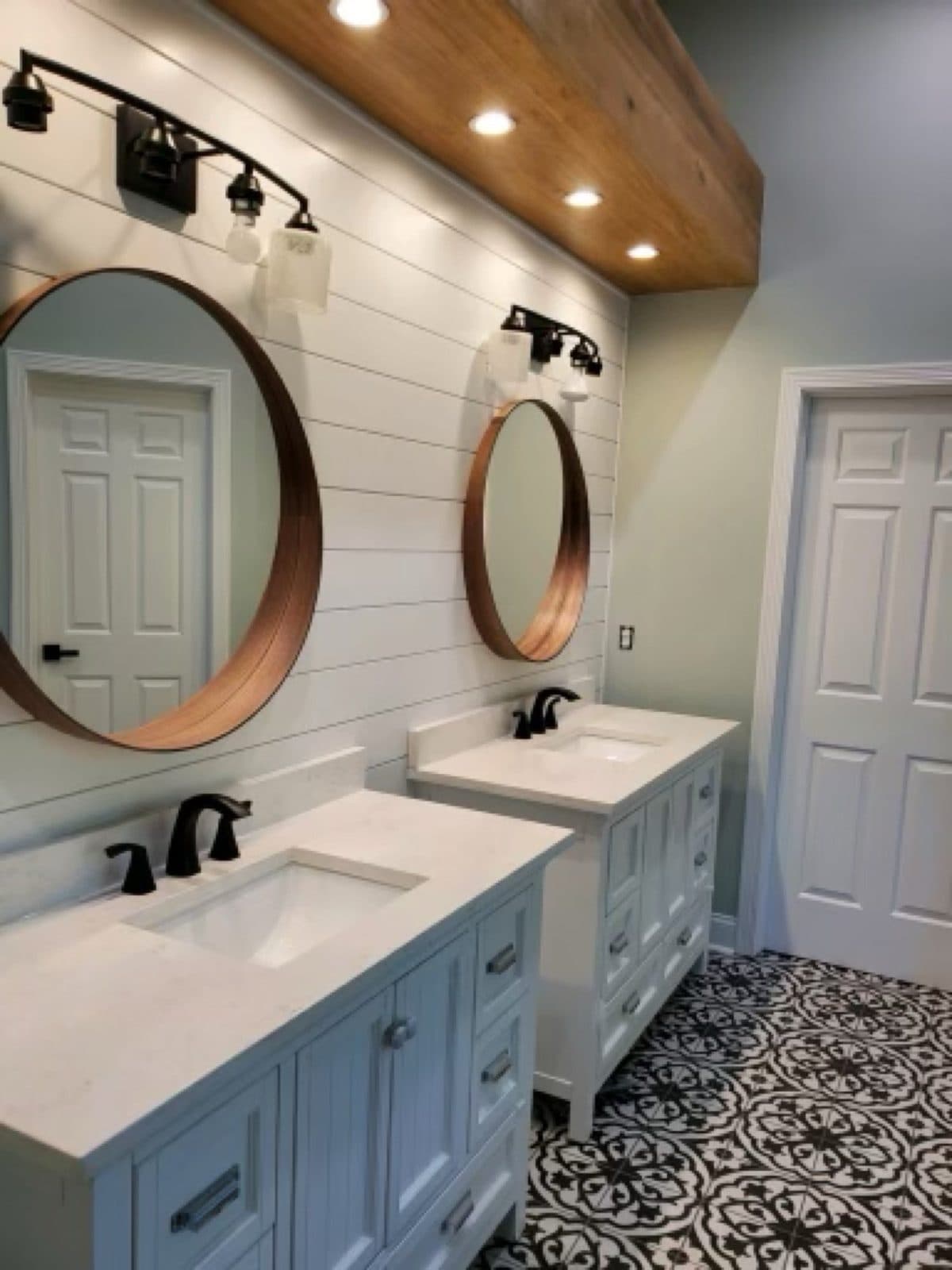 A double vanity under a shiplap wall, two round brass mirrors, black faucets, and a patterned black-and-white tile floor.
