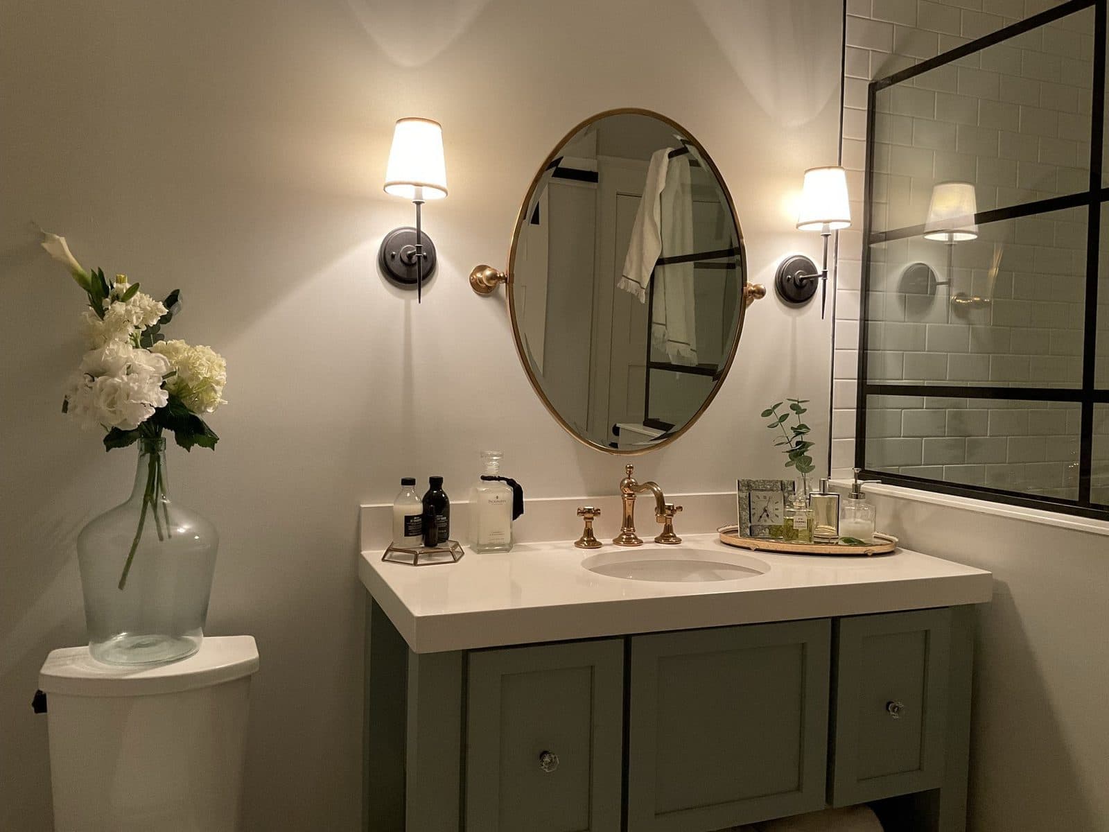 The powder room: a round mirror and brass sconces over a sage-blue vanity, a glass vase of white blooms on the counter.