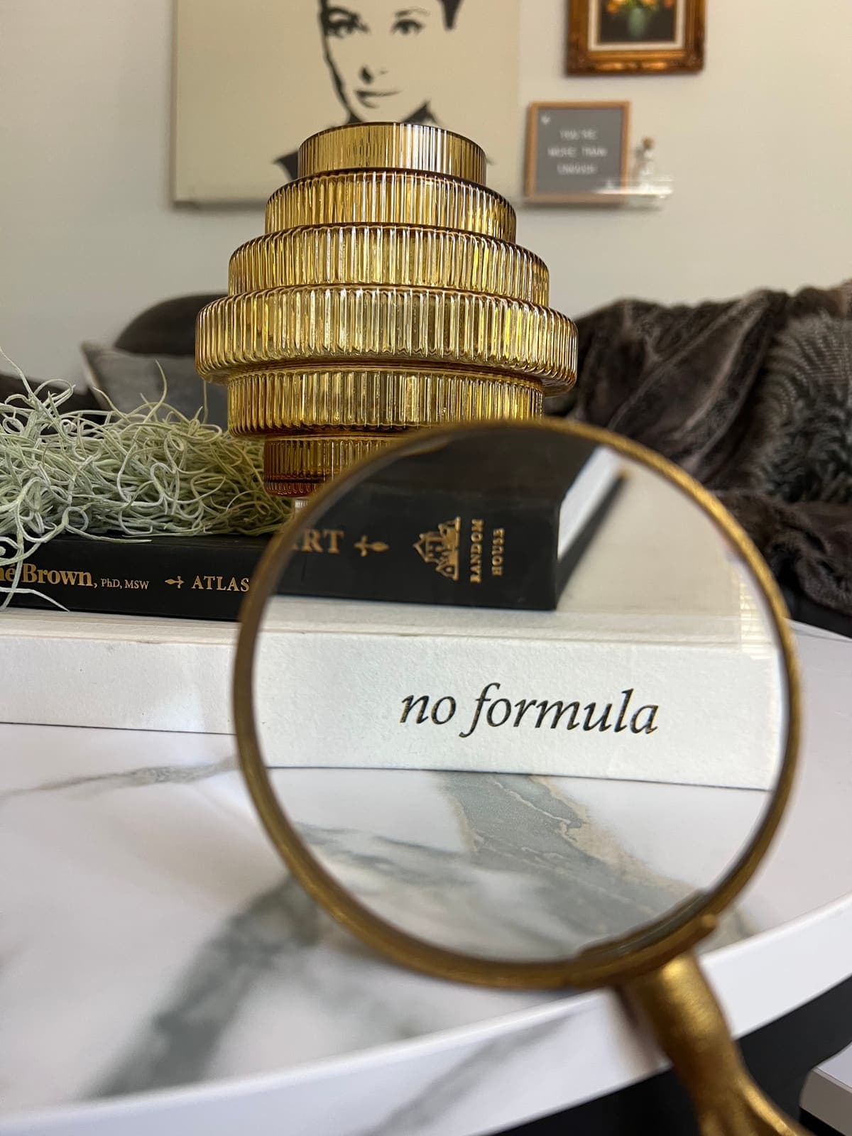 A magnifying glass resting over the words 'no formula' on a book, on a marble tabletop beside a ribbed brass lamp.