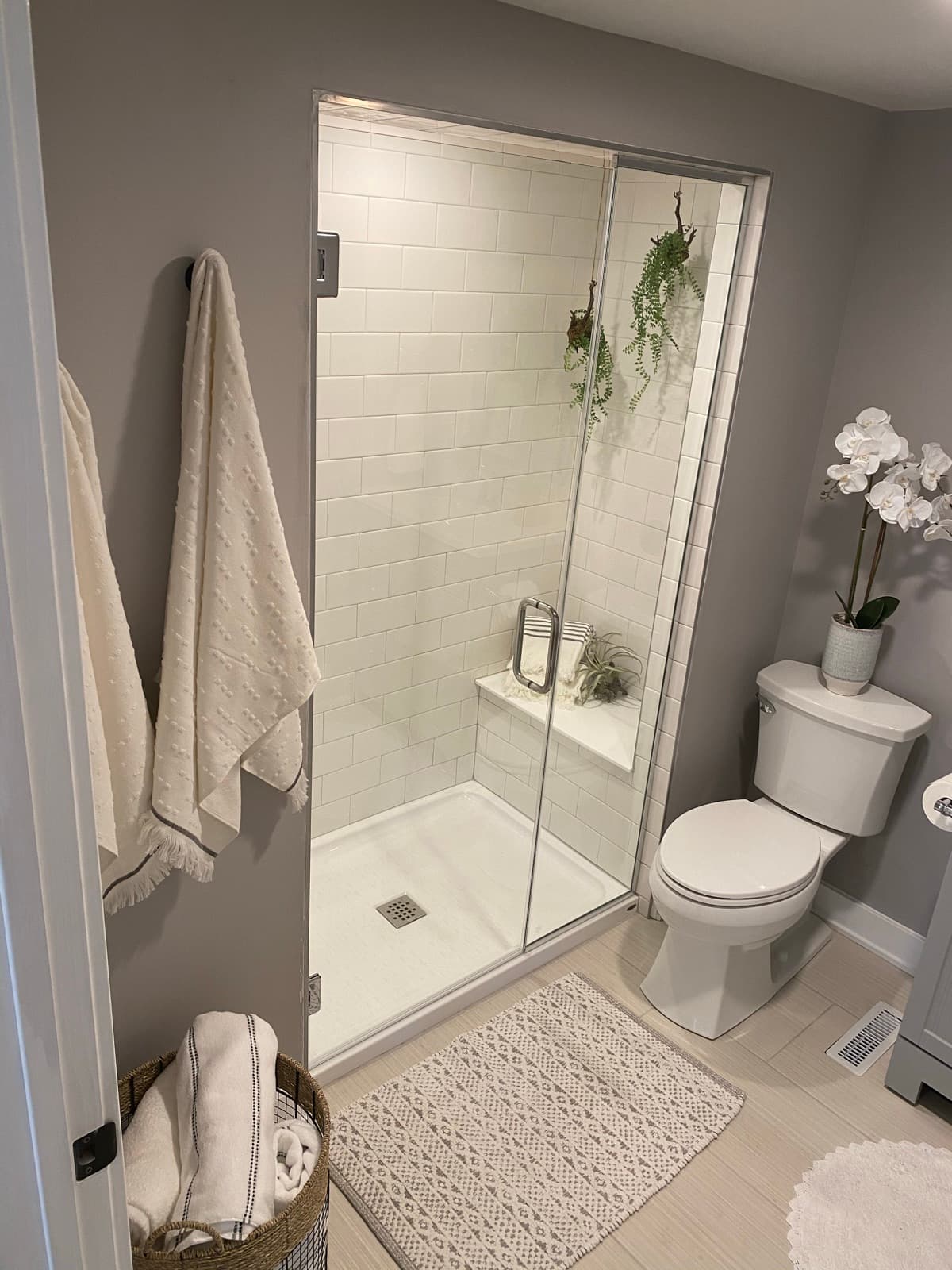 Spa-styled bath, glass shower with trailing greenery, plush towels, orchid, and a patterned mat.