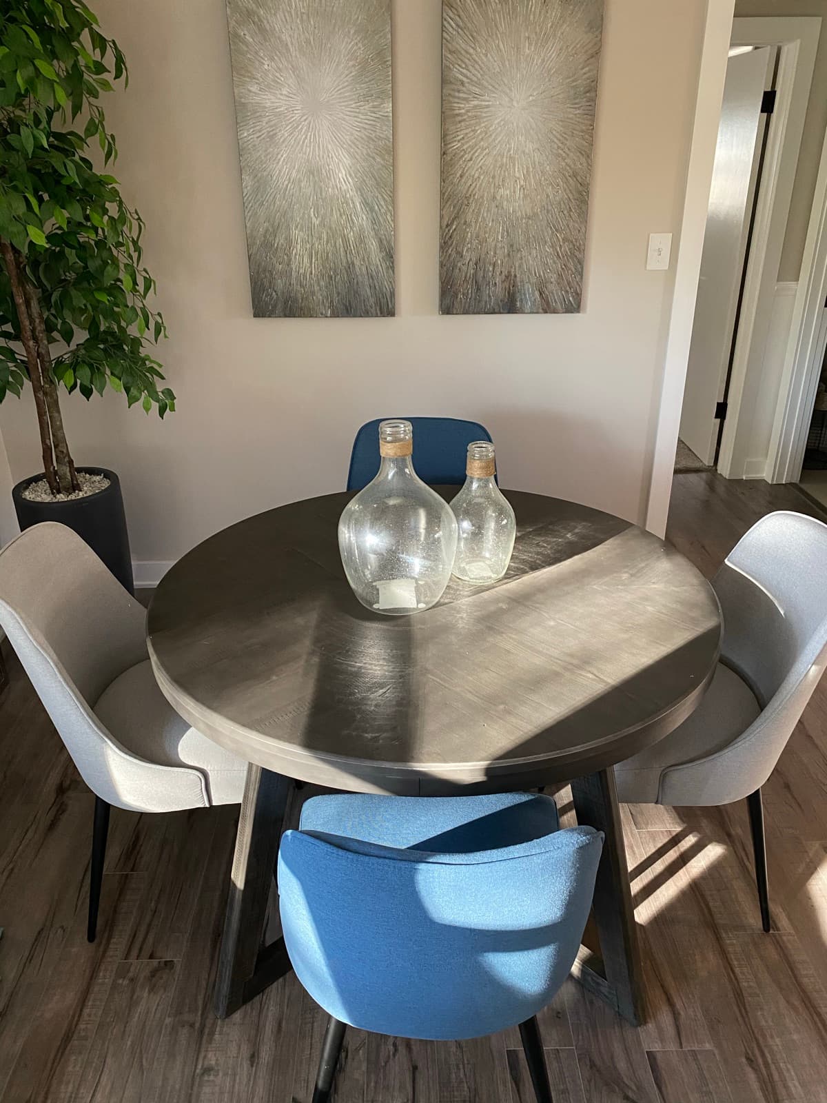 Round wood dining table with mixed upholstered chairs, glass demijohns, and a pair of starburst canvases.