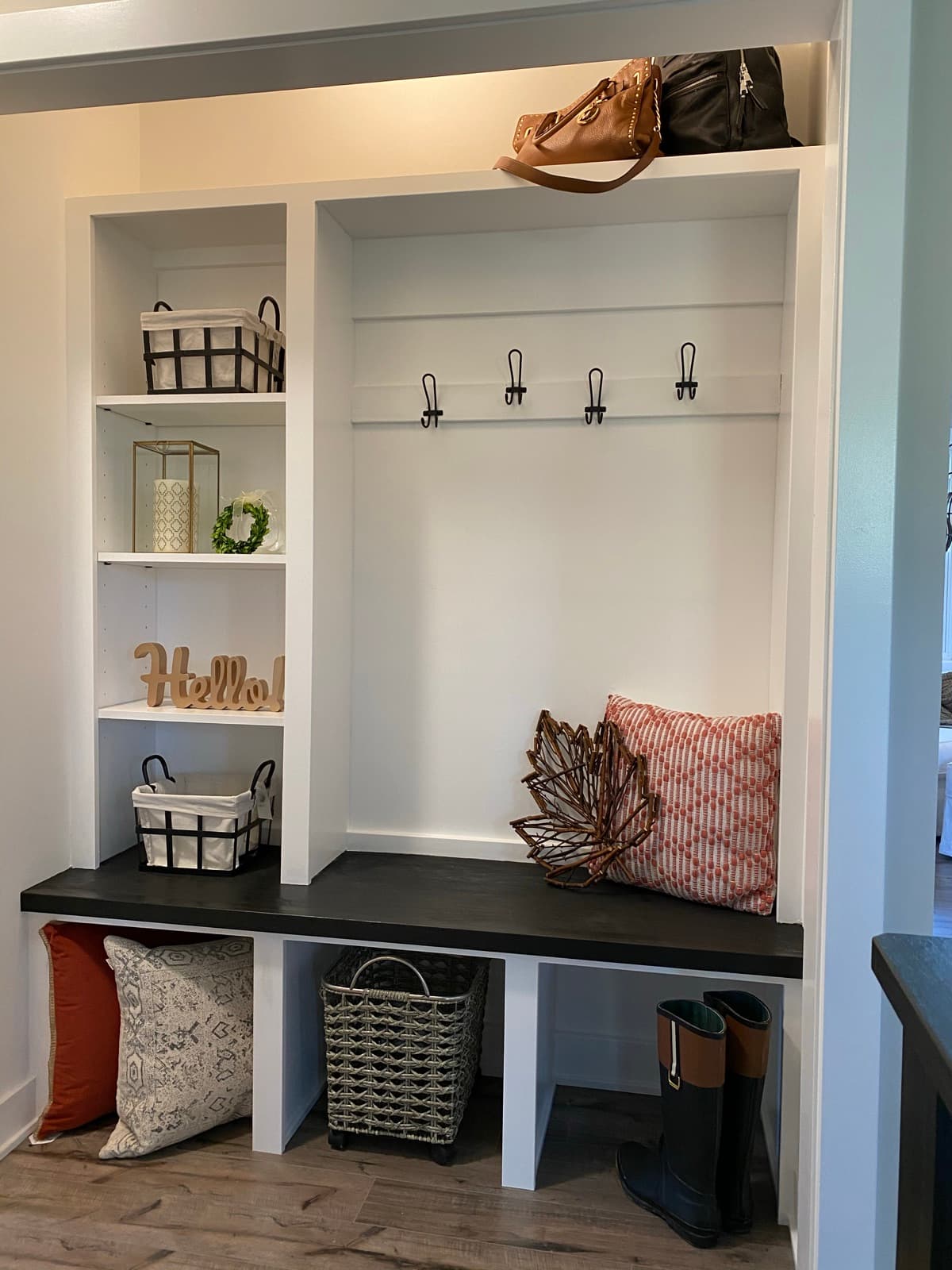Built-in drop zone styled with baskets, a 'Hello' sign, rust pillows, and a bench beneath coat hooks.