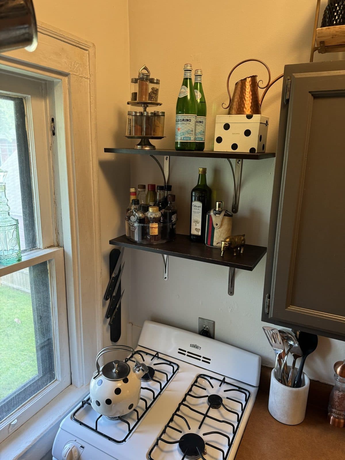 A polka-dot kettle on the stove under open shelves of bottles and copper, the window letting the garden in.