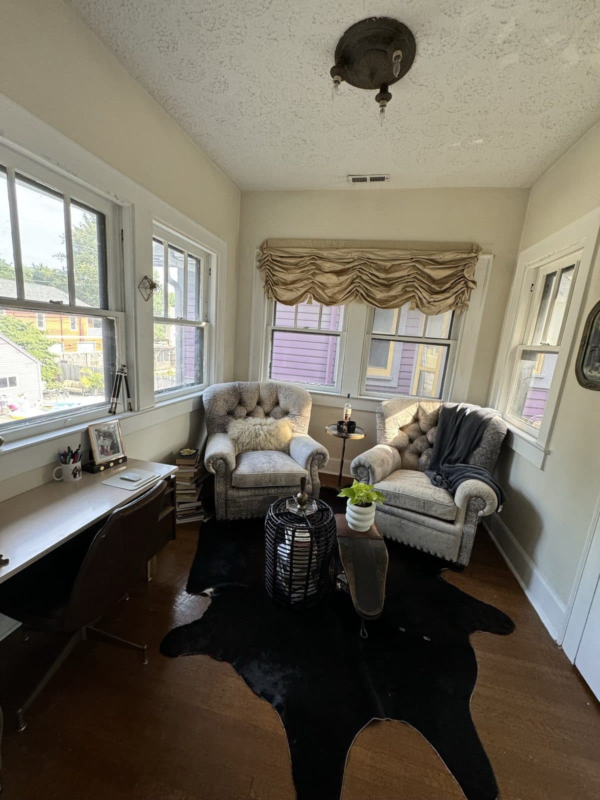 The sunroom: two tufted grey armchairs, a black rattan side table, and a cowhide rug in front of a wall of windows.