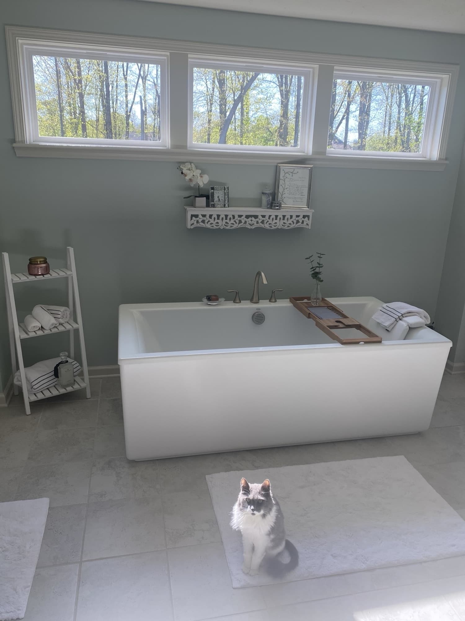 The tub and window bank from the side, a ladder shelf of rolled towels alongside, styled with candles and a wall shelf of frames and orchid.