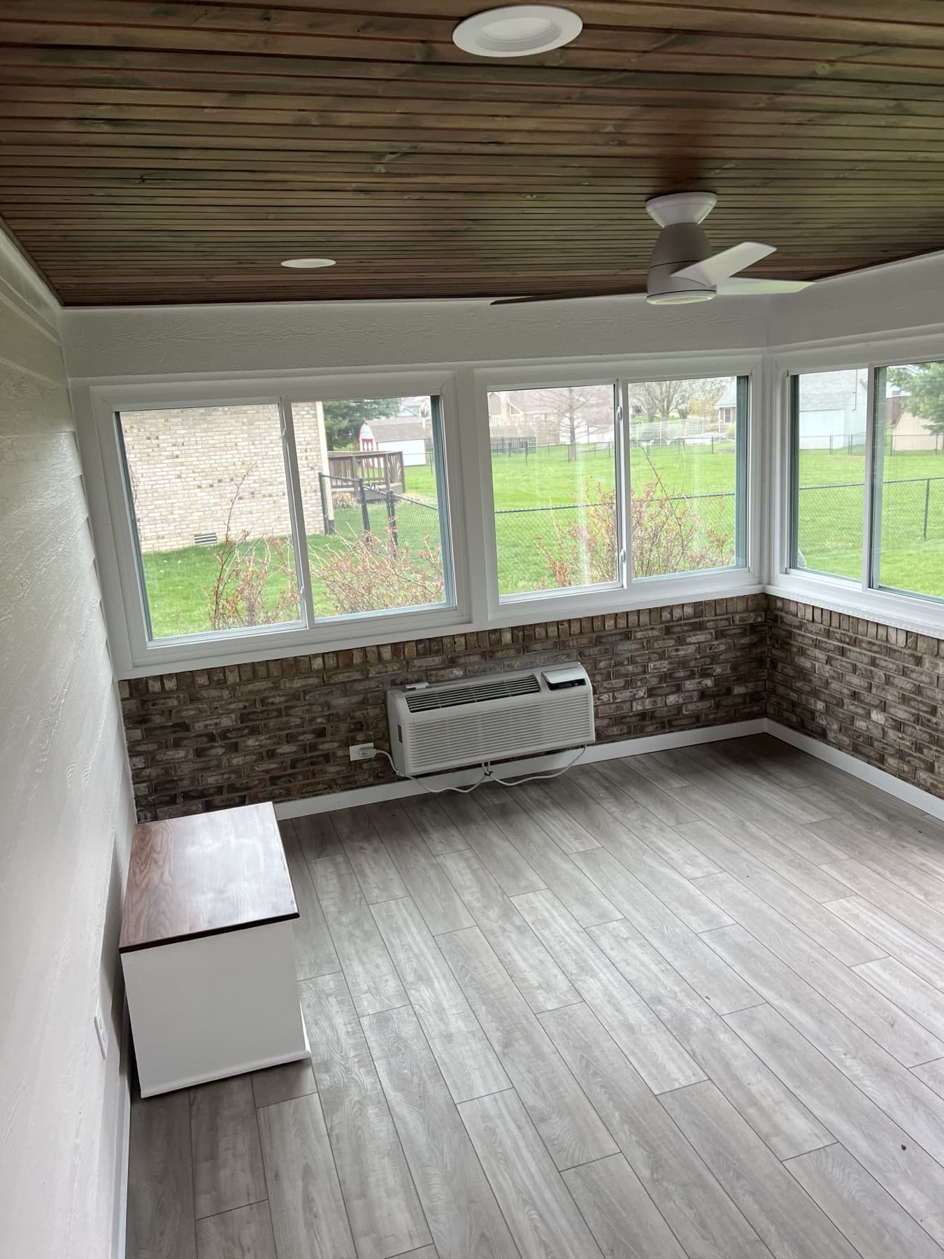 The sunroom before styling, empty with a gray wood-look floor, wood-plank ceiling, and brick knee wall.